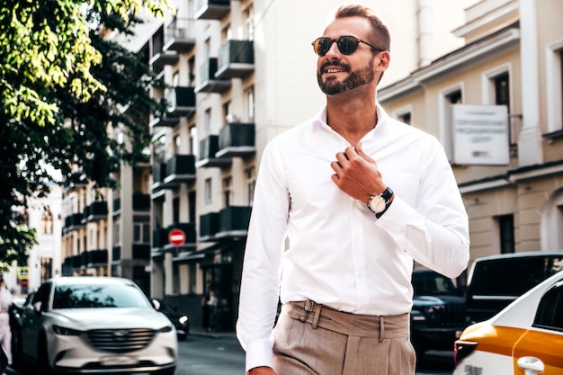 Retrato de guapo modelo lambersexual hipster con estilo seguro Hombre moderno sexy vestido con camisa blanca y pantalones Hombre de moda posando en el fondo de la calle en la ciudad de Europa al atardecer Con gafas de sol