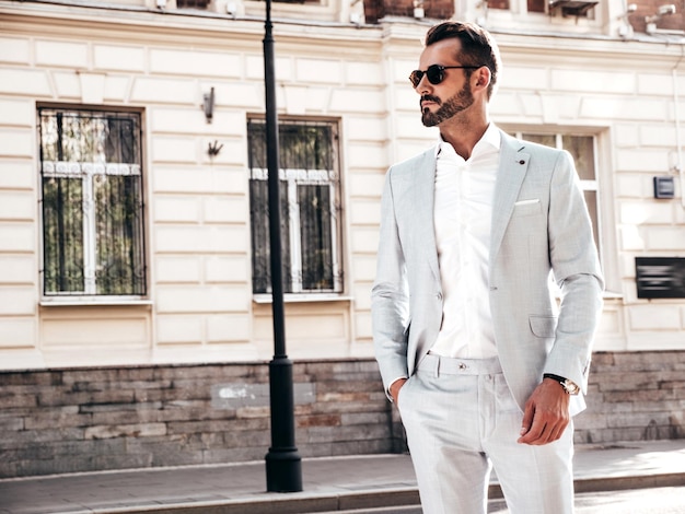 Retrato de guapo modelo lambersexual hipster con estilo seguro Hombre moderno sexy vestido con elegante traje blanco Hombre de moda posando en el fondo de la calle en la ciudad de Europa al atardecer Con gafas de sol