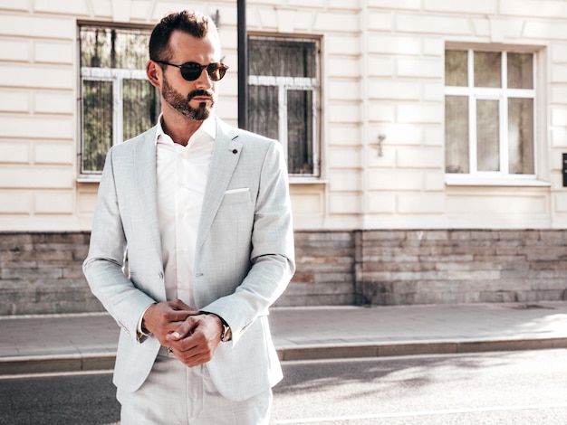 Retrato de guapo modelo lambersexual hipster con estilo seguro Hombre moderno sexy vestido con elegante traje blanco Hombre de moda posando en el fondo de la calle en la ciudad de Europa al atardecer Con gafas de sol