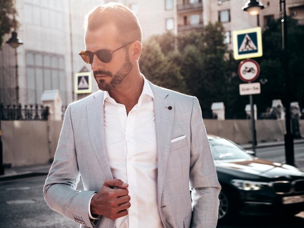 Retrato de guapo modelo lambersexual hipster con estilo seguro Hombre moderno sexy vestido con elegante traje blanco Hombre de moda posando en el fondo de la calle en la ciudad de Europa al atardecer Con gafas de sol