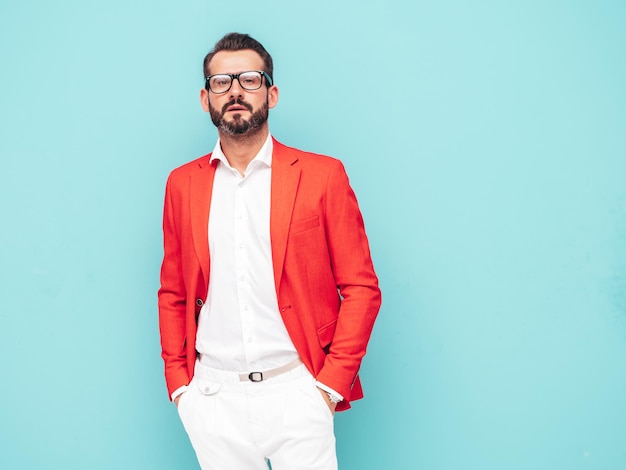 Retrato de guapo modelo lambersexual hipster con estilo seguro Hombre moderno sexy vestido con elegante traje rojo Hombre de moda posando en el estudio cerca de la pared azul en espectáculos