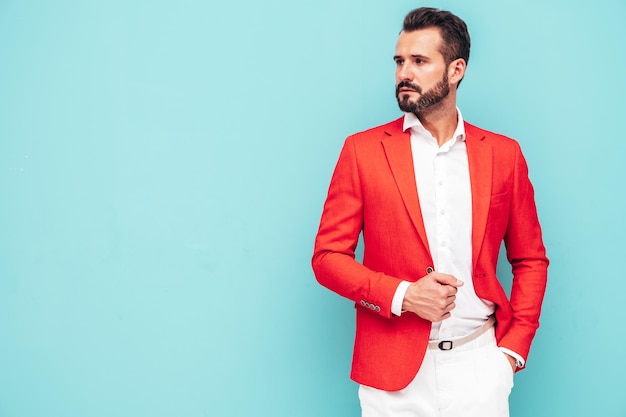 Retrato de guapo modelo lambersexual hipster con estilo seguro Hombre moderno sexy vestido con elegante traje rojo Hombre de moda posando en el estudio cerca de la pared azul