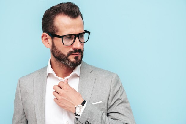 Retrato de guapo modelo lambersexual hipster con estilo seguro Hombre moderno sexy vestido con traje elegante y espectáculos Hombre de moda posando en el estudio cerca de la pared azul Mirando a la cámara con gafas