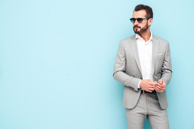 Retrato de guapo modelo lambersexual hipster con estilo seguro Hombre moderno sexy vestido con traje elegante Hombre de moda posando en el estudio cerca de la pared azul con gafas de sol