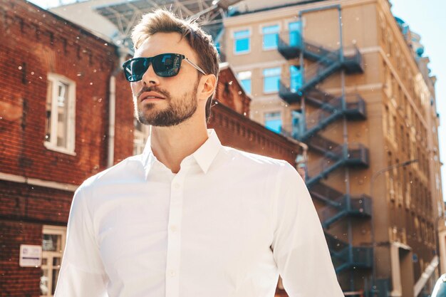 Retrato de guapo modelo lambersexual hipster con estilo seguro Hombre moderno vestido con camisa blanca Hombre de moda posando en el fondo de la calle cerca de rascacielos con gafas de sol Al aire libre al atardecer