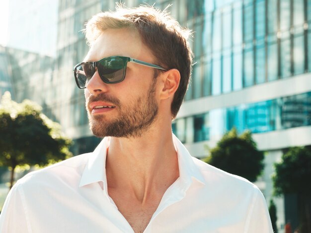 Retrato de guapo modelo lambersexual hipster con estilo seguro Hombre moderno vestido con camisa blanca Hombre de moda posando en el fondo de la calle cerca de rascacielos con gafas de sol Al aire libre al atardecer