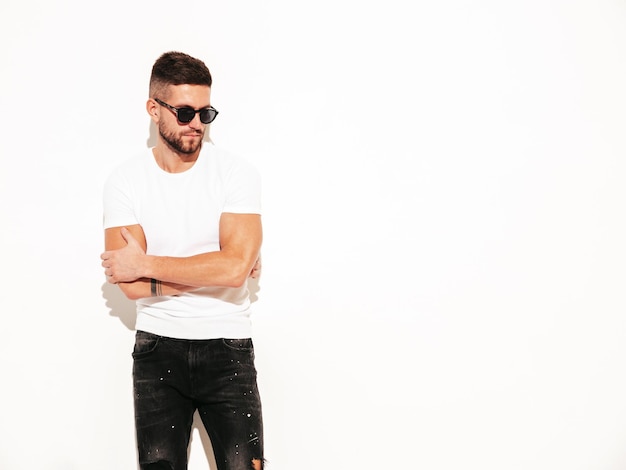 Retrato de guapo modelo lambersexual hipster con estilo seguro hombre vestido con camiseta blanca y jeans hombre de moda posando cerca de la pared en el estudio con gafas de sol