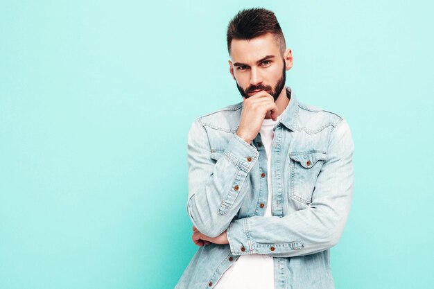 Retrato de guapo modelo lambersexual hipster con estilo seguro Hombre vestido con chaqueta y jeans Hombre de moda aislado en la pared azul en el estudio Pensativo tocando su barbilla