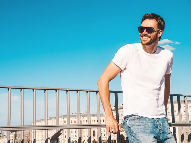 Retrato de guapo modelo lambersexual hipster sonriente Hombre elegante vestido con camiseta blanca Hombre de moda posando detrás del cielo azul en el fondo de la calle con gafas de sol