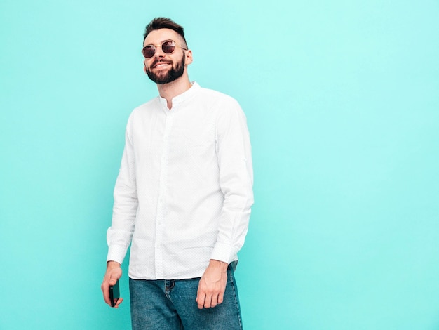 Foto gratuita retrato de guapo modelo sonriente hombre elegante sexy vestido con camisa blanca y jeans hombre hipster de moda posando junto a la pared azul en el estudio aislado en gafas de sol