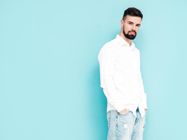 Foto gratuita retrato de guapo modelo sonriente hombre elegante sexy vestido con camisa blanca y jeans hombre hipster de moda posando junto a la pared azul en el estudio aislado