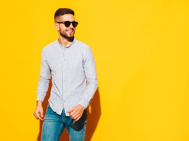 Retrato de guapo modelo sonriente Hombre elegante sexy vestido con camisa y jeans Hombre hipster de moda posando junto a la pared amarilla en el estudio con gafas de sol Verano