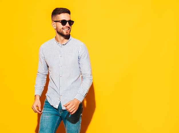 Retrato de guapo modelo sonriente Hombre elegante sexy vestido con camisa y jeans Hombre hipster de moda posando junto a la pared amarilla en el estudio con gafas de sol Verano
