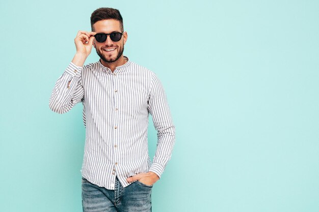 Retrato de guapo modelo sonriente Hombre elegante sexy vestido con camisa y jeans Hombre hipster de moda posando junto a la pared azul en el estudio Alegre y feliz En gafas de sol