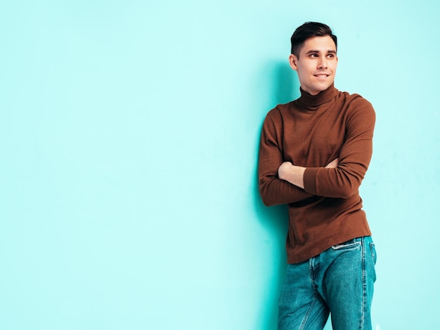 Retrato de guapo modelo sonriente Hombre elegante sexy vestido con suéter de cuello alto y jeans Hombre hipster de moda posando cerca de la pared azul en el estudio Brazos cruzados aislados