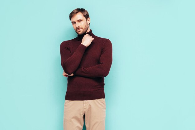 Retrato de guapo modelo sonriente Hombre elegante sexy vestido con suéter de cuello alto y pantalones Hombre hipster de moda posando junto a la pared azul en el estudio Aislado