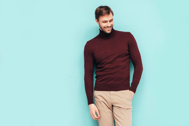 Retrato de guapo modelo sonriente Hombre elegante sexy vestido con suéter de cuello alto y pantalones Hombre hipster de moda posando junto a la pared azul en el estudio Aislado