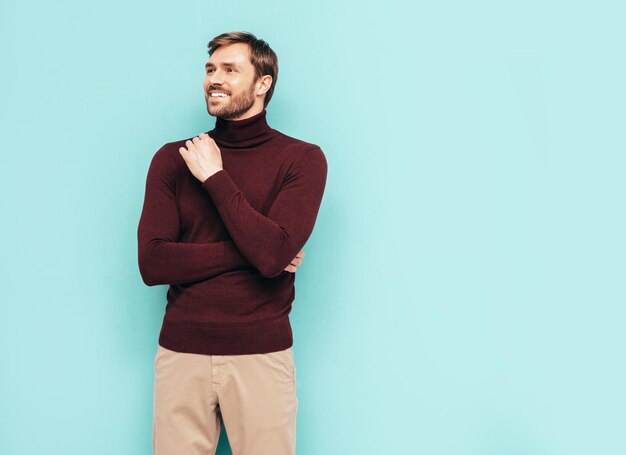 Retrato de guapo modelo sonriente Hombre elegante sexy vestido con suéter de cuello alto y pantalones Hombre hipster de moda posando junto a la pared azul en el estudio Aislado