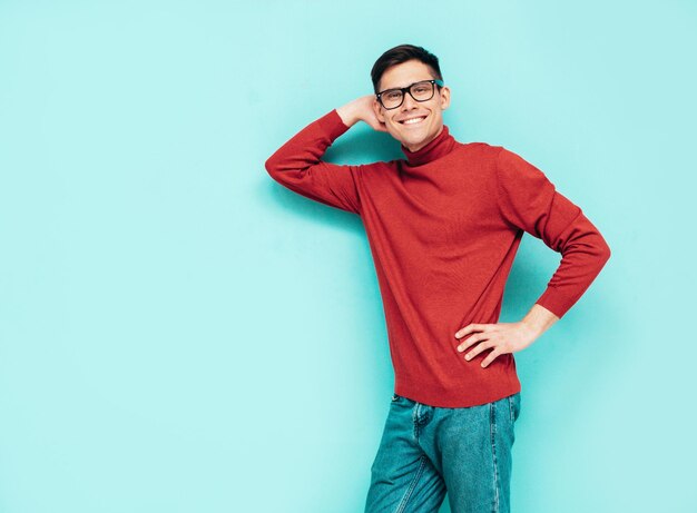Retrato de guapo modelo sonriente Hombre elegante sexy vestido con suéter de cuello alto rojo y jeans Hombre hipster de moda posando junto a la pared azul en el estudio Aislado en gafas de gafas