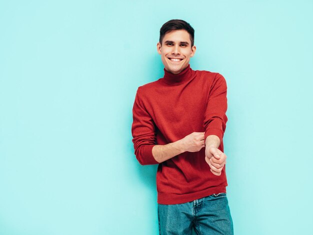 Retrato de guapo modelo sonriente Hombre elegante sexy vestido con suéter de cuello alto rojo y jeans Hombre hipster de moda posando junto a la pared azul en el estudio Aislado
