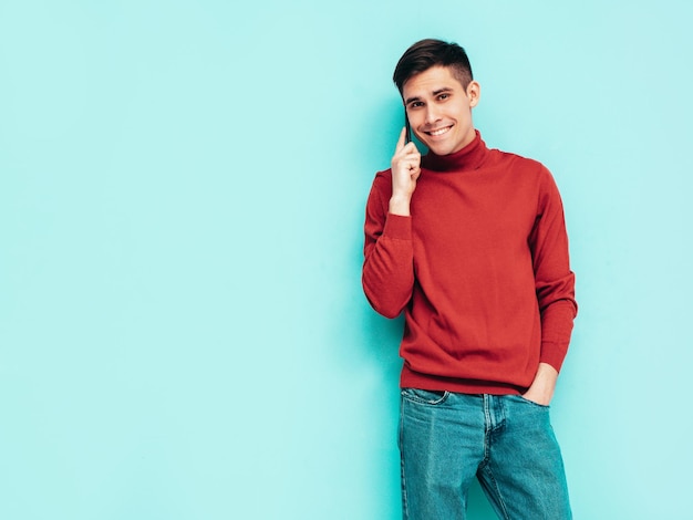 Retrato de guapo modelo sonriente Hombre elegante sexy vestido con suéter de cuello alto rojo y jeans Hombre hipster de moda posando junto a la pared azul en el estudio Sosteniendo teléfono Con teléfono celular aislado