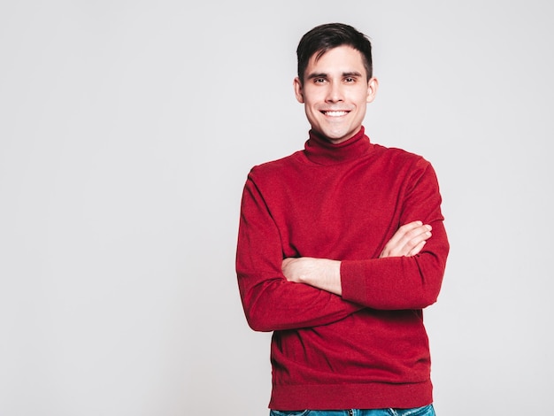 Retrato de guapo modelo sonriente Hombre elegante sexy vestido con suéter de cuello alto rojo y jeans Hombre hipster de moda posando sobre fondo gris en estudio Brazos cruzados aislados