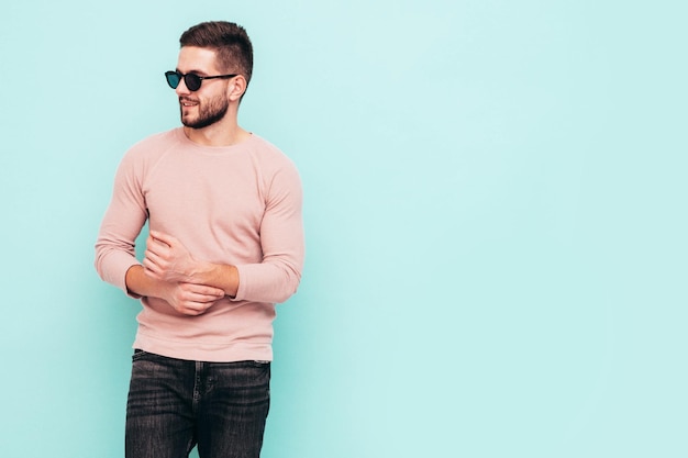 Retrato de guapo modelo sonriente Hombre elegante sexy vestido con suéter rosa y jeans Hombre hipster de moda posando junto a la pared azul en el estudio En gafas de sol Aislado