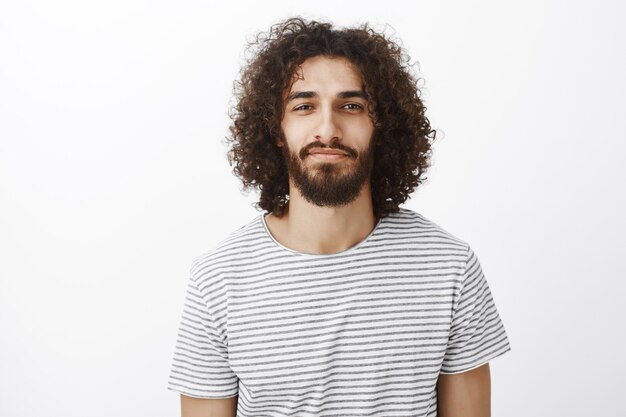 Retrato de guapo novio creativo seguro con cabello oscuro y rizado, de pie con una leve sonrisa y expresión segura de sí mismo