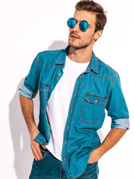Retrato de guapo sonriente elegante joven modelo vestido con ropa de jeans. Hombre de moda con gafas de sol.