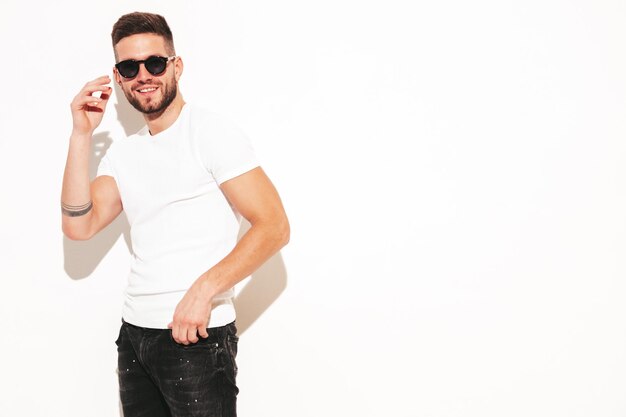 Retrato de guapo sonriente con estilo hipster lambersexual modelMan vestido con camiseta blanca y jeans Hombre de moda posando cerca de la pared en el estudio con gafas de sol