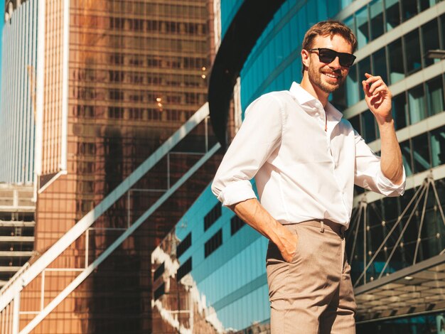 Retrato de guapo sonriente con estilo hipster lambersexual modelModern hombre vestido con camisa blanca Hombre de moda posando en el fondo de la calle cerca de rascacielos con gafas de sol