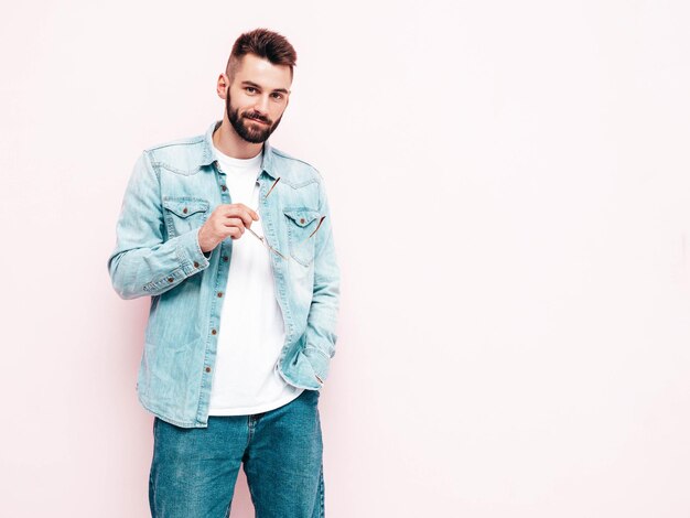 Retrato de guapo sonriente con estilo hipster lambersexual modelo Hombre vestido con chaqueta y jeans Hombre de moda aislado en la pared rosa en el estudio