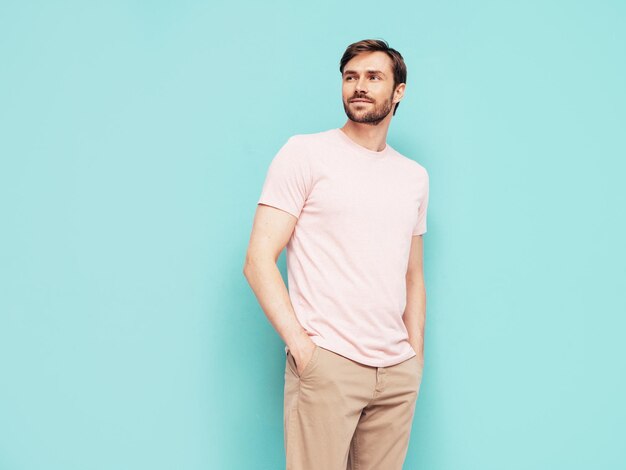 Retrato de guapo sonriente con estilo hipster lambersexual modelo Sexy hombre vestido con camiseta rosa y pantalones Hombre de moda aislado en la pared azul en el estudio