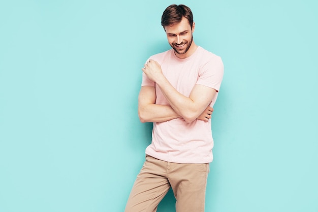 Retrato de guapo sonriente con estilo hipster lambersexual modelo Sexy hombre vestido con camiseta rosa y pantalones Hombre de moda aislado en la pared azul en el estudio