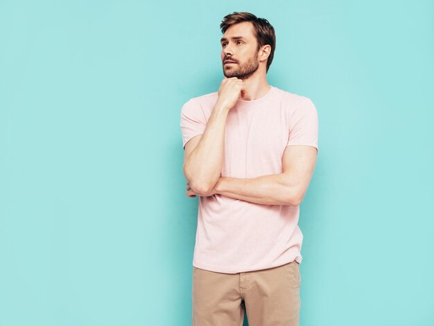 Retrato de guapo sonriente con estilo hipster lambersexual modelo Sexy hombre vestido con camiseta rosa y pantalones Hombre de moda aislado en la pared azul en el estudio