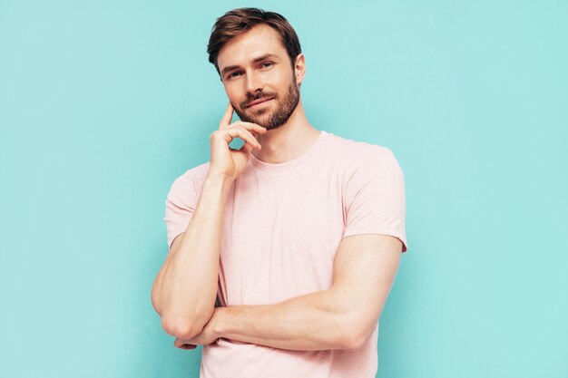 Retrato de guapo sonriente con estilo hipster lambersexual modelo Sexy hombre vestido con camiseta rosa y pantalones Hombre de moda aislado en la pared azul en el estudio