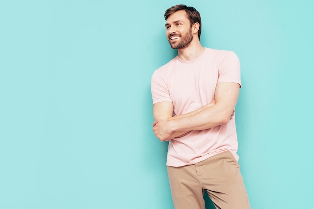 Retrato de guapo sonriente con estilo hipster lambersexual modelo Sexy hombre vestido con camiseta rosa y pantalones Hombre de moda aislado en la pared azul en el estudio