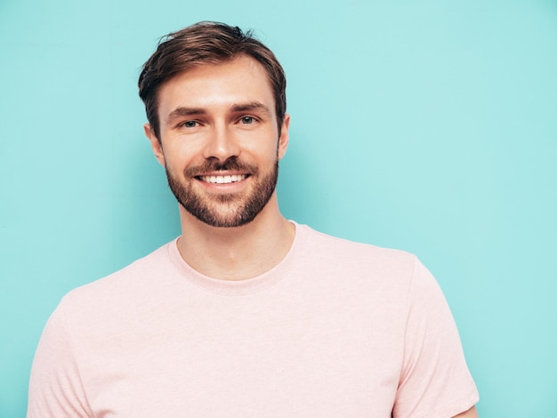 Retrato de guapo sonriente con estilo hipster lambersexual modelo Sexy hombre vestido con camiseta rosa y pantalones Hombre de moda aislado en la pared azul en el estudio