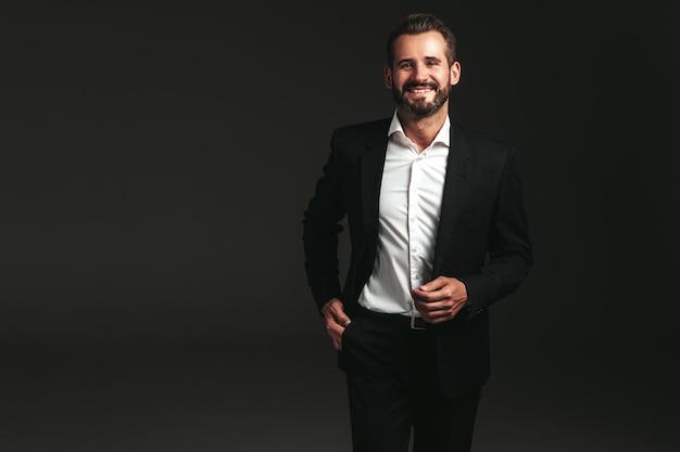 Retrato de guapo sonriente con estilo hipster lambersexual modelo Sexy moderno hombre vestido con elegante traje negro Moda hombre posando en estudio sobre fondo oscuro Mirando a la cámara