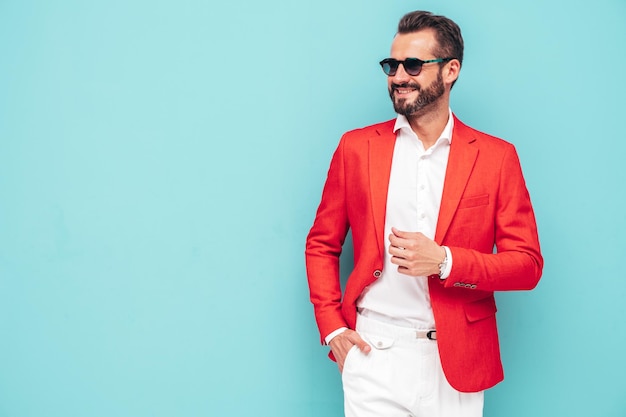 Retrato de guapo sonriente con estilo hipster lambersexual modelSexy hombre moderno vestido con elegante traje rojo Hombre de moda posando en el estudio cerca de la pared azul en gafas de sol