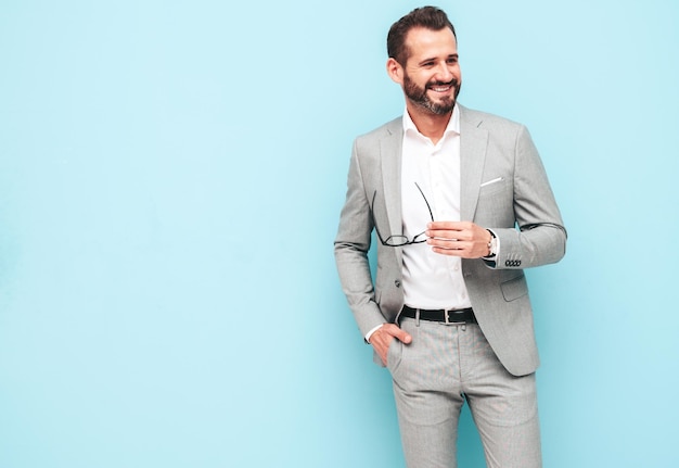 Retrato de guapo sonriente con estilo hipster lambersexual modelSexy hombre moderno vestido con traje elegante Hombre de moda posando en el estudio cerca de la pared azul