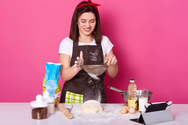 Retrato de hábil y talentoso cocinero poniendo harina a través del tamiz en medio pastel listo con pasas. Morena linda joven modelo plantea aislado en rosa brillante. Concepto de cocción y horneado.