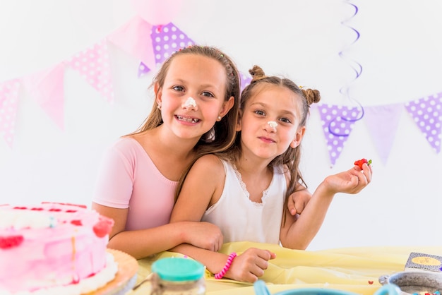 Retrato de hermanas lindas con pastel en la nariz disfrutando en fiesta de cumpleaños
