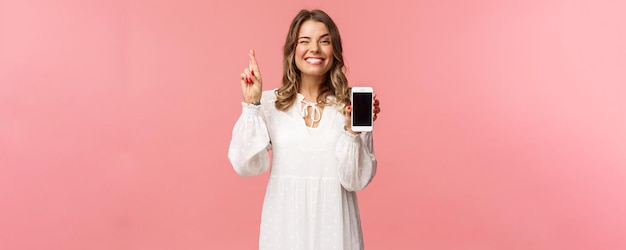 Retrato de una hermosa y afortunada chica rubia con vestido blanco haciendo apuestas mostrando la pantalla del teléfono móvil con el dedo cruzado buena suerte y sonriendo optimista, tenga fe en que ganará en la competencia