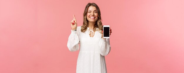 Retrato de una hermosa y afortunada chica rubia con vestido blanco haciendo apuestas mostrando la pantalla del teléfono móvil con el dedo cruzado buena suerte y sonriendo optimista, tenga fe en que ganará en la competencia