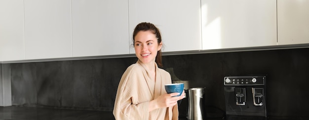 Foto gratuita retrato de una hermosa ama de casa mujer joven en bata de baño sosteniendo una taza de café bebiendo té en el