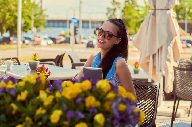 El retrato de una hermosa bloguera feliz que usa ropa de moda y gafas de sol sostiene una tableta que disfruta del día de verano mientras se sienta en una terraza en un café al aire libre, mirando la cámara.