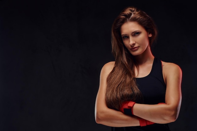 Retrato de una hermosa boxeadora deportiva con ropa deportiva y manos vendadas, posando con los brazos cruzados. Aislado en un fondo de textura oscura.