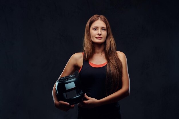 El retrato de una hermosa boxeadora morena con ropa deportiva sostiene un casco protector, mirando una cámara. Aislado en un fondo de textura oscura.