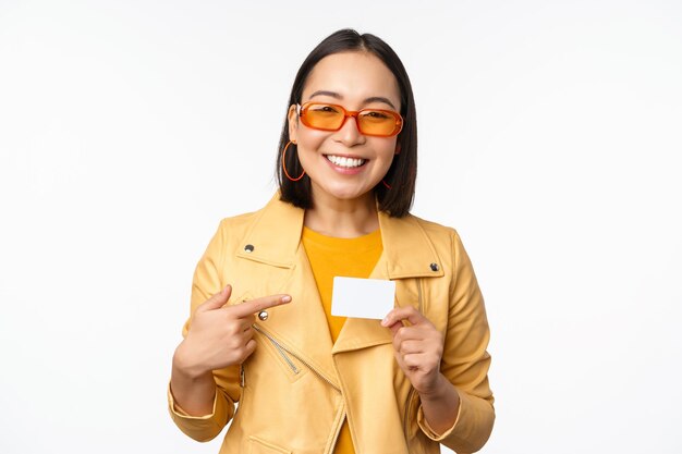 Retrato de una hermosa chica asiática moderna con gafas de sol sonriendo feliz mostrando una tarjeta de crédito de pie sobre un espacio de copia de fondo blanco